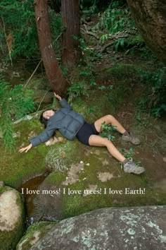 a woman laying on top of a moss covered rock next to a forest filled with trees