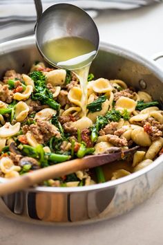 a skillet filled with pasta, sausage and broccoli being poured into it