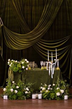 an arrangement of flowers and candles on a table in front of a green drape