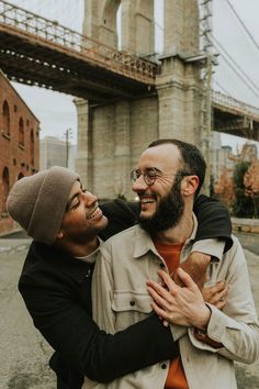 two men are hugging each other in front of the brooklyn bridge, one is wearing glasses