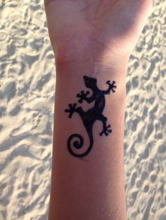 a small lizard tattoo on the wrist of a person's hand, with sand in the background