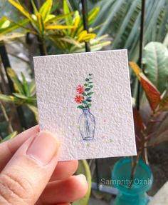 a hand holding up a small card with flowers in a glass jar on top of it