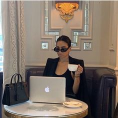 a woman sitting at a table in front of a laptop computer holding a cup of coffee