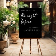 a welcome sign in front of a building with potted plants on the sidewalk and flowers behind it