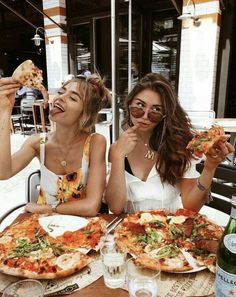 two beautiful women sitting at a table with pizza and drinks in front of their faces