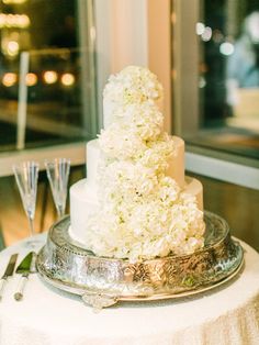 a wedding cake with white flowers on top sits on a silver platter next to wine glasses