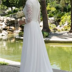 a woman standing in front of a pond wearing a white wedding dress with long sleeves