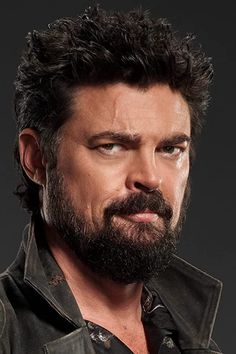 a man with black hair and beard wearing a leather shirt looking at the camera while standing in front of a dark background