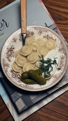 a white plate topped with ravioli and pickles next to a knife on top of a magazine