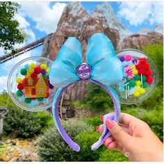 a person holding up a mickey mouse ears headband with balloons in front of a mountain