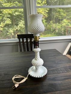 a white lamp sitting on top of a wooden table next to a window with trees in the background