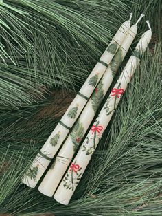 three white candles with holly and bows on them sitting next to some pine needles in the grass