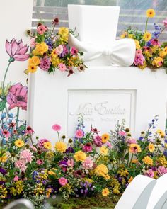 a white sign with flowers around it and a bow tie on top