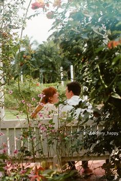 a man and woman sitting on a bench in the middle of some bushes with pink flowers