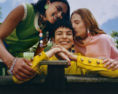 three young women leaning on a wooden bench with their arms around each other and smiling at the camera