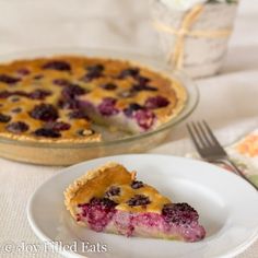 a slice of blueberry pie on a white plate with a fork and glass vase