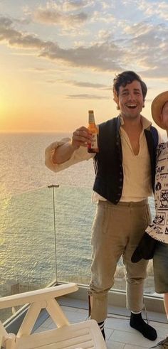 two men standing next to each other on top of a roof near the ocean at sunset