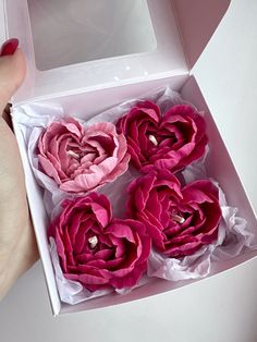 four pink flowers in a white box on a table with a person's hand