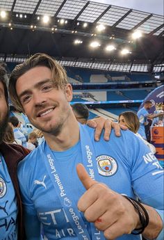 two men in blue shirts giving the thumbs up at a soccer game with fans behind them