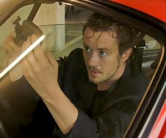 a man sitting in the driver seat of a red car holding up a cell phone