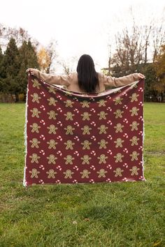 a woman holding up a red and gold christmas themed blanket in the grass with her back to the camera