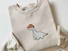 Embroidered Mushroom Dinosaur Sweatshirt, Mushroom Dinosaur Shirt, Paleontology Shirt Dino Shirt Velociraptor Shirt Trex Shirt Dinosaur Gift, CozyGenZ This Cute Embroidered Mushroom Dinosaur Sweatshirt is a must have for Dinosaur lovers! What's better than looking cute and warm in the colder months? Our crewnecks are made from the highest quality fabric for an incredibly soft and comfortable fit, with advanced printing and embroidery to ensure that the colors and graphics are vibrant and detailed. ❤ If you want to order matching shirts, add each item separately by choosing different colors and sizes each time, and add each one into your cart. ❤ Sizes run from Small to XXL (2XL). This unisex sweater is intended to be oversized and loose-fitting for women for a chic and laid-back vibe. If yo Mushroom Dinosaur, Embroidered Christmas Gifts, Dinosaur Sweatshirt, Dino Shirt, Dinosaur Gifts, Dinosaur Shirt, Personalized Embroidery, Dinosaur Christmas, Custom Sweatshirts