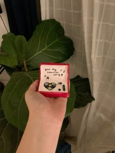 a hand holding a small red and white box with writing on it next to a large green plant