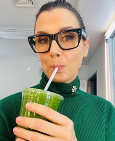 a woman wearing glasses drinking a green smoothie with a straw in her mouth and looking at the camera