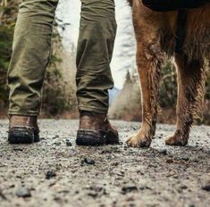 two people standing next to a brown dog on a dirt road with trees in the background