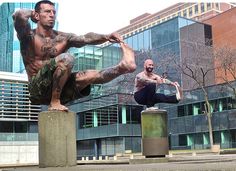 two men are doing yoga outside in front of a large building and one is standing on his legs