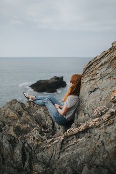 a woman sitting on top of a rocky cliff next to the ocean with her legs crossed