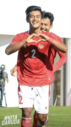 two young men standing next to each other on top of a soccer field with their hands in the shape of a heart