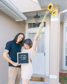 a man and woman holding up a sunflower in front of a house with a sign