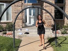 a woman standing in front of a house with an arch on the sidewalk and potted plants behind her