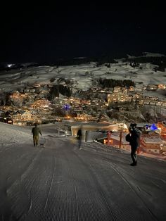 two people walking up the side of a snow covered slope at night with city lights in the background