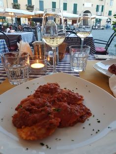 a white plate topped with meat covered in sauce and garnish next to glasses of wine