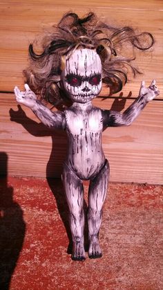 a creepy doll with long hair standing on the ground in front of a wooden wall