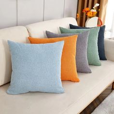 four different colored pillows on a white couch in front of a vase with orange flowers