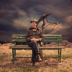 a man sitting on top of a green bench next to a dead tree in the middle of a field