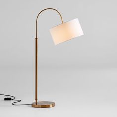 a floor lamp with a white shade on the top and a gold base, in front of a gray background