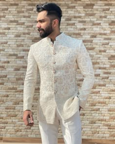 a man standing in front of a brick wall wearing white pants and a jacket with an intricate pattern