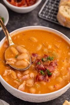 a bowl of soup with beans and bacon in it, next to bread on the side