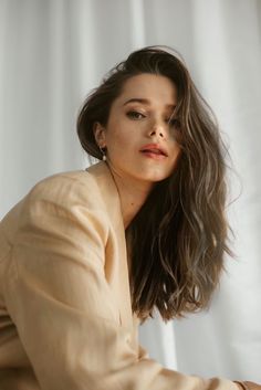 a woman with long hair sitting on a chair in front of a white curtain and looking off to the side