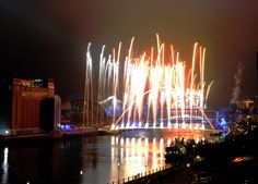 fireworks are lit up in the night sky over a river with buildings and lights on it