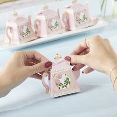 two hands holding a pink teapot shaped ornament on a table with plates and napkins in the background