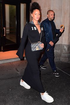 a woman is walking down the street in a black dress and white tennis shoes with her handbag