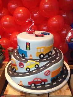 a three tiered cake with cars and balloons in the background at a birthday party