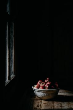 a bowl full of strawberries sitting on a table next to a window in a dark room