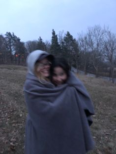 two women wrapped in a blanket are smiling at the camera while standing in a field