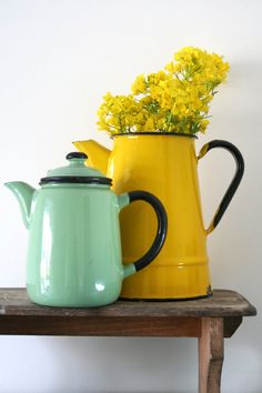 two teapots with yellow flowers in them sitting on a wooden table next to a white wall